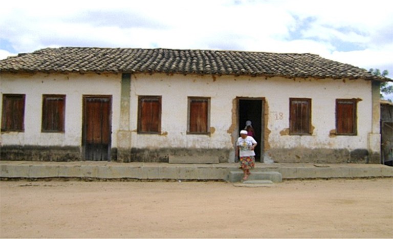 A imagem mostra uma casa antiga em estilo colonial rústico ao fundo, uma mulher sai na porta de turbante, camiseta e saia.